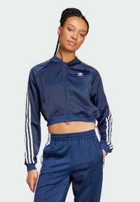 Navy blue cropped athletic jacket in smooth fabric with white stripes along sleeves, featuring zip closure and small logo on chest.