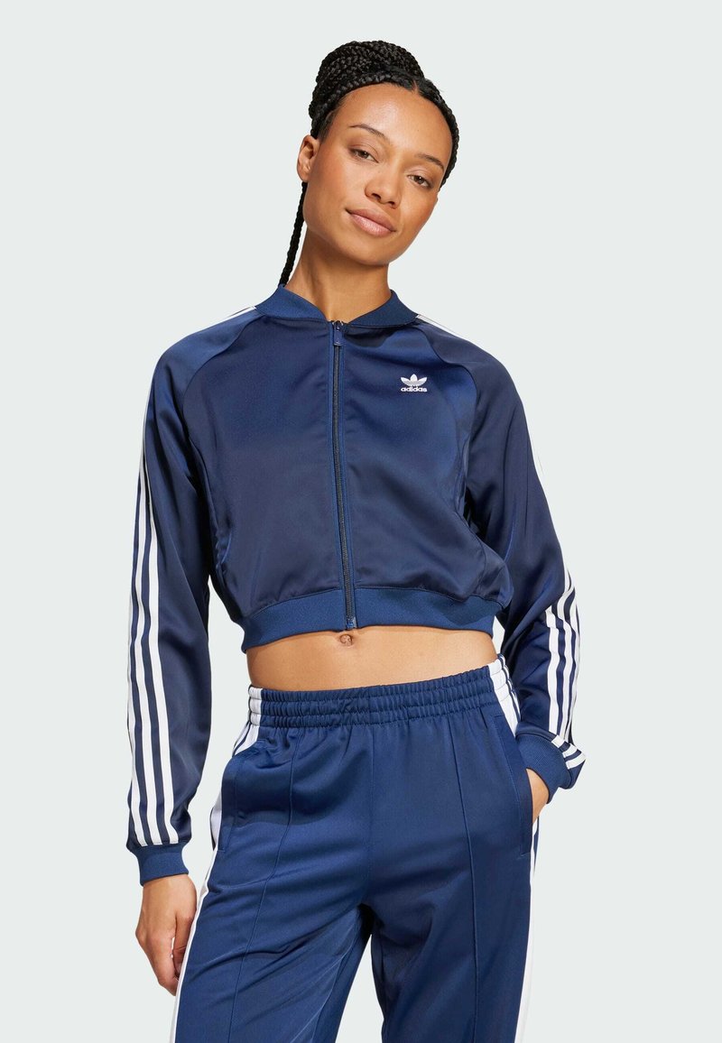 Navy blue cropped athletic jacket in smooth fabric with white stripes along sleeves, featuring zip closure and small logo on chest.