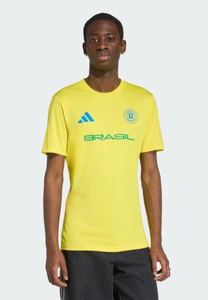Ung mand iført en gul brasiliansk sportstrøje med blå logoer og sorte bukser, stående mod en ensfarvet lys baggrund.