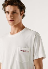Camiseta blanca de algodón con cuello redondo y mangas cortas, con texto bordado en rojo en el bolsillo izquierdo del pecho.