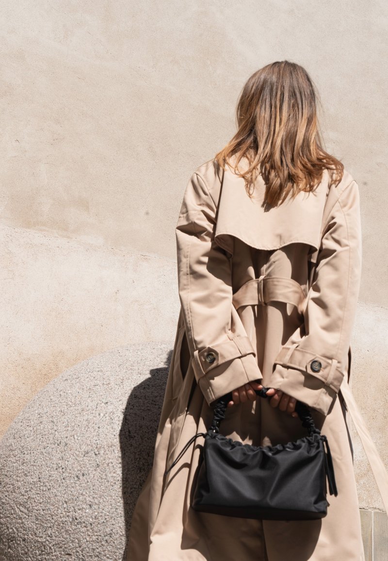 Mujer con cabello hasta los hombros, vistiendo un abrigo beige y sosteniendo un bolso negro detrás de la espalda, de pie contra una pared beige.