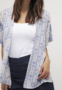 Lätt mönstrad kimono med blå och vita ränder samt blommönster. Kort ärm, öppen framtill, bärs över vitt linne och mörk kjol.