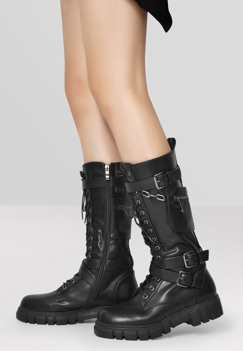 Botas de cuero negras hasta la rodilla con cordones en la parte frontal, cremallera y múltiples correas. Suela de goma gruesa con detalles de metal.