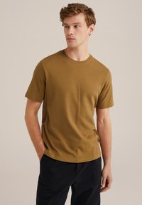 Kaki spalvos trumpomis rankovėmis marškinėliai iš minkštos medvilnės, su apvalia iškirpte. Atsitiktinis kirpimas su lygia tekstūra, derinami su tamsiomis kelnėmis.