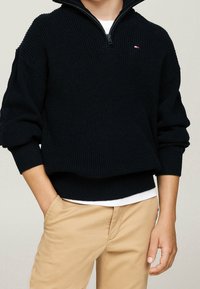 Pull noir en maille avec zip, col montant, présentant une texture côtelée et un petit logo sur la poitrine, porté par-dessus une chemise blanche.