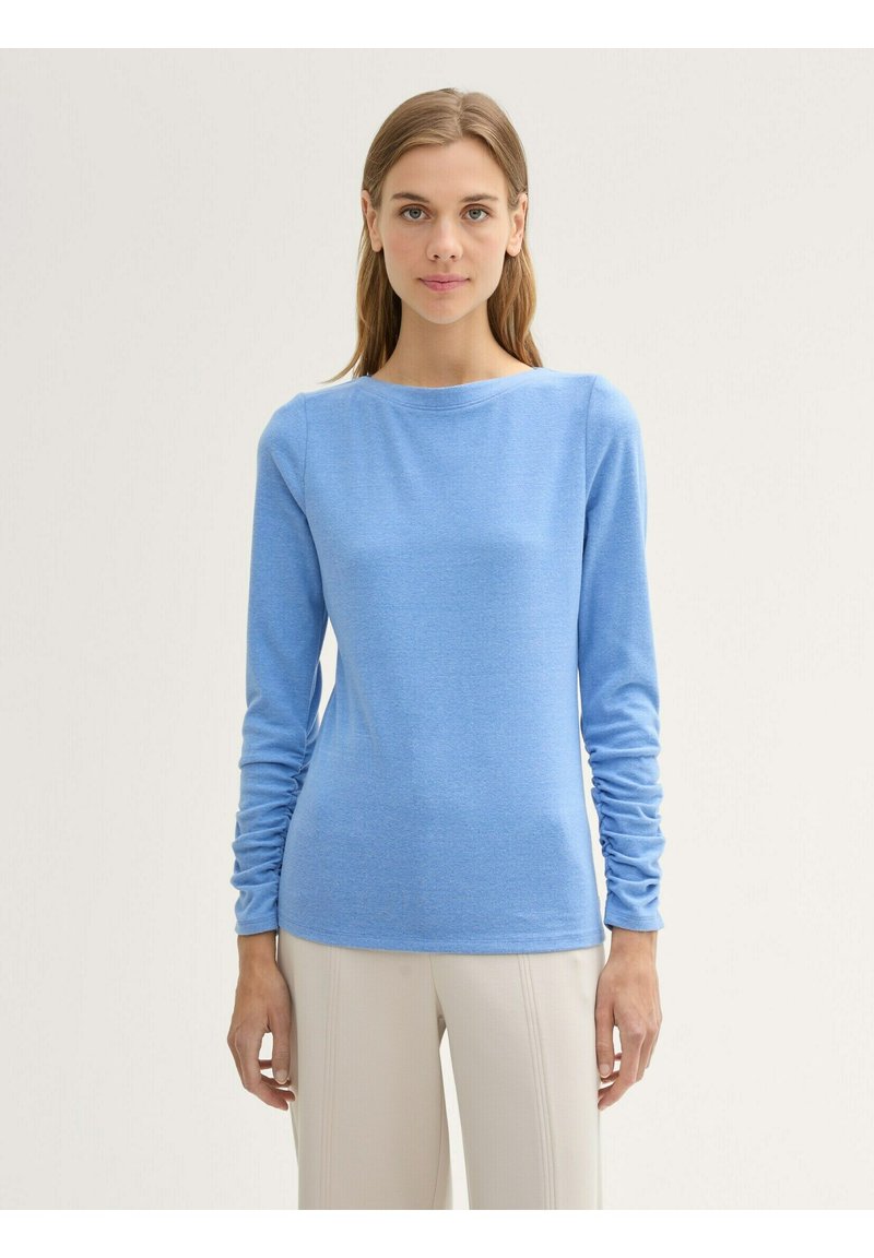 Maglietta azzurra chiara a maniche lunghe con scollo rotondo, maniche arricciate e una texture liscia. Indossata con pantaloni color crema.