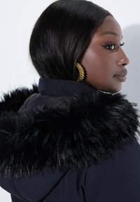 Chaqueta navy con un borde de capucha de piel sintética negra. Presenta una textura suave y un diseño de cuello alto. Pendiente de aro dorado visible.
