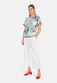 Kurzärmlige Bluse aus hellblauem Stoff mit bunten abstrakten Mustern, kombiniert mit weißen weit geschnittenen Hosen und roten Blockabsatzschuhen.