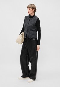 Hallide tekstuuriga hall ilma varrukateta vest nupuga, must pikkade varrukatega särk all, must nahast laiad püksid ja beež kott.