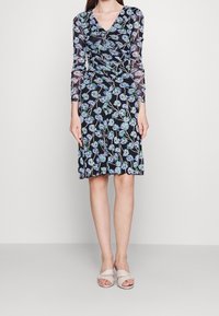 Schwarzes Blumenkleid mit V-Ausschnitt, langen durchsichtigen Ärmeln, floralen Mustern in Blau und Grün und einem ausgestellten Rock. Kombiniert mit beigen Riemchensandalen.