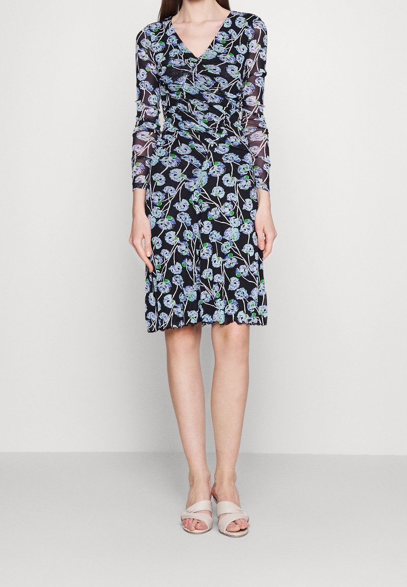 Schwarzes Blumenkleid mit V-Ausschnitt, langen durchsichtigen Ärmeln, floralen Mustern in Blau und Grün und einem ausgestellten Rock. Kombiniert mit beigen Riemchensandalen.