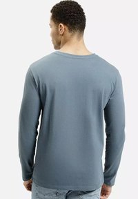 Mężczyzna z krótkimi kręconymi włosami, ubrany w prostą niebieską koszulę z długim rękawem i jasnoniebieskie dżinsy, stojący tyłem na białym tle.