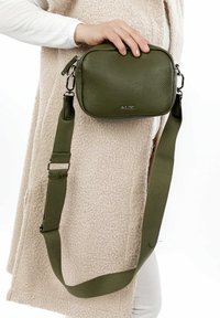 Borsa a tracolla in similpelle verde oliva con forma quadrata, finitura testurizzata, accessori in metallo e tracolla in tessuto regolabile.