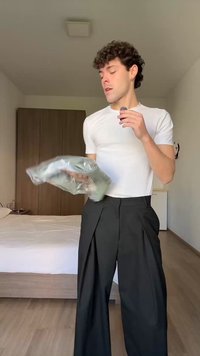 Mlada osoba u bijeloj majici i širokim crnim hlačama drži presavijenu odjeću u plastičnoj vrećici dok stoji u spavaćoj sobi.