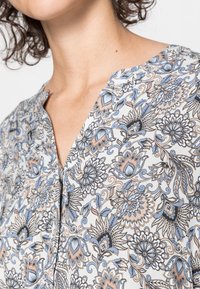 Donna che indossa una blusa bianca con motivi floreali blu e beige, visibile dalla zona del collo fino alla parte superiore del petto e della spalla.
