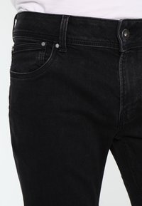 Schwarze Jeans aus Denim mit klassischem Schnitt, fünf Taschen und metallischen Designelementen am Bund. Glatte Textur mit leichten Verwaschungen.