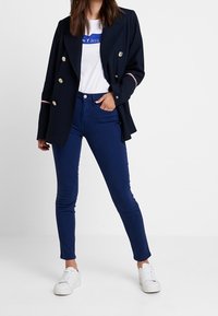 Blazer azul marino de doble botonadura con botones dorados sobre una camiseta blanca con letras azules, combinado con jeans ajustados azul oscuro y zapatillas blancas.
