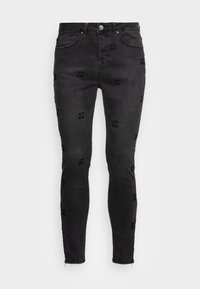 Black denim skinny jeans featuring embossed text patterns, five pockets, and a zippered ankle detail. Classic cut and sturdy fabric.