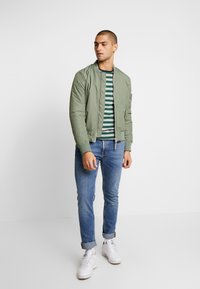 Chaqueta bomber verde oliva con puños acanalados, llevada sobre una camisa de rayas azul marino y blanca, combinada con jeans de mezclilla azul claro y zapatillas blancas.