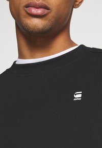 Sudadera de algodón negra con cuello redondo, con un cuello interior blanco y un pequeño logo "RAW" blanco en el pecho. Textura suave.