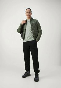 Mannen gevoerde jas in donkergroen, met ritssluiting, gedragen over een lichtgroene t-shirt. Zwarte broek en contrasterende zwarte sneakers maken de outfit compleet.