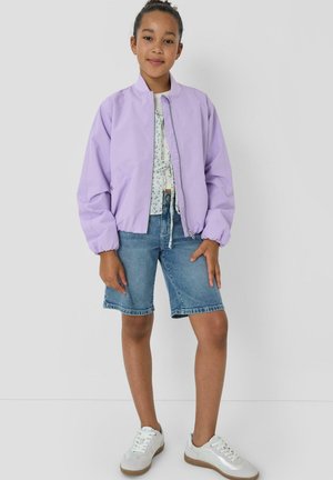 Dziewczyna stojąca w jasnofioletowej kurtce zapinanej na zamek, kwiecistej bluzce, dżinsowych spodenkach do kolan i białych trampkach na jednolitym tle.