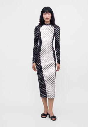 Femme aux longs cheveux noirs porte une robe midi ajustée noire et blanche avec de petits motifs en forme de cœur et des sandales à talons noires.