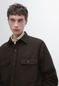 Bruine wolblend overhemdjack met een knoopsluiting, twee borstzakken en een klassieke kraag, gecombineerd met een lichtgekleurde blouse eronder.