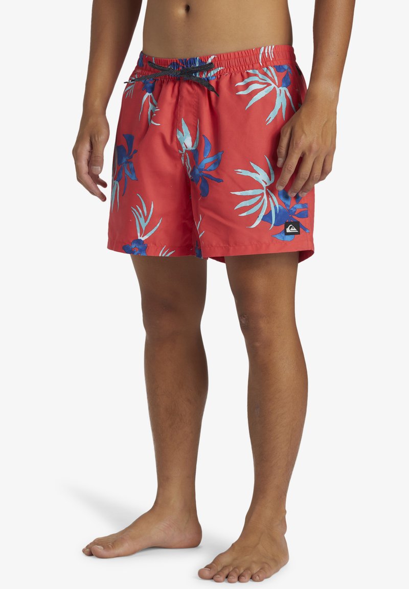 Bañador rojo con un patrón de hojas tropicales en azul y verde azulado, cintura elástica con cordón y parche de logo en la esquina inferior izquierda.