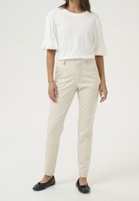 Beżowe spodnie o dopasowanym kroju, z przednimi kieszeniami i gładkim wykończeniem, zestawione z białą bluzką z plisowanymi rękawami oraz czarnymi płaskimi butami.