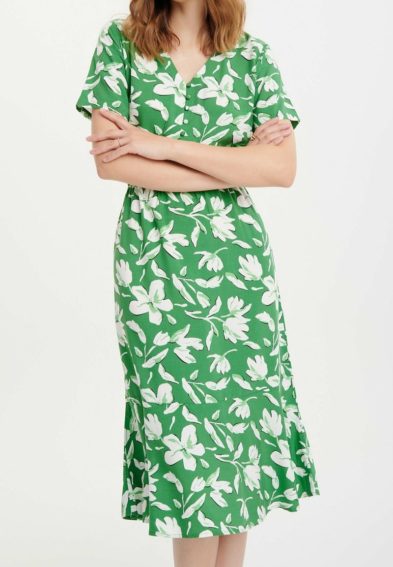 Grön klänning med vitt blommönster, korta ärmar, V-ringning och utsvängd fåll. Materialet verkar lätt och andas.