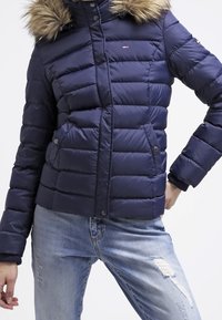 Žena oblečená v tmavě modré prošívané bundě s kapucí lemovanou kožešinou a světle modrých roztrhaných džínách, s rukama na boky.