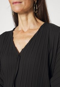 Svart plisserad klänning med V-halsdesign. Har texturerat tyg och draperade ärmar. Fästa statementörhängen med geometriska inslag.