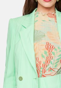 Light green double-breasted blazer with subtle vertical stripes, paired with a patterned blouse featuring orange and green floral designs.