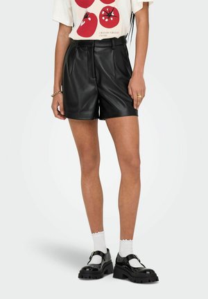 Frau trägt schwarze Leder-Shorts, weißes T-Shirt mit rotem Tomatendruck, weiße Socken und schwarze klobige Mary-Jane-Schuhe.