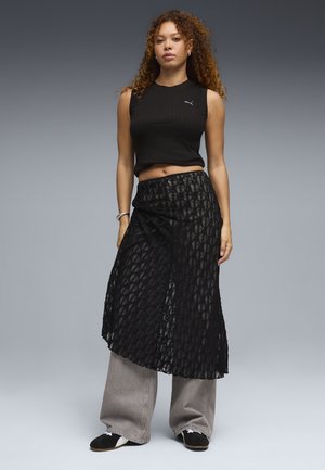 Vrouw met krullend haar, gekleed in een mouwloos zwart cropped topje, een gestructureerde zwarte wikkelrok over brede grijze broek, en zwarte sneakers, staat stil.