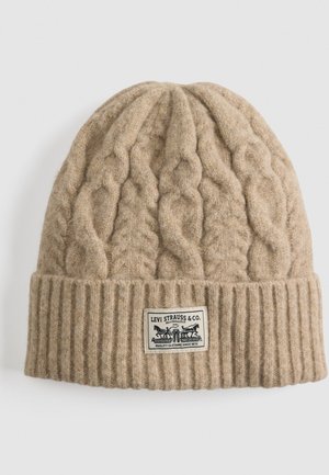 Bonnet en tricot beige avec un revers côtelé, présentant un design en tricot torsadé. Inclut une étiquette rectangulaire avec la marque "Levi Strauss & Co.".