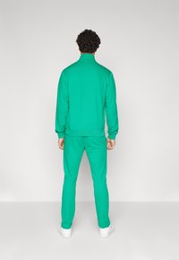 Green tracksuit with high collar, fitted cuffs, and matching pants. Soft texture and elastic waistband, paired with white sneakers.