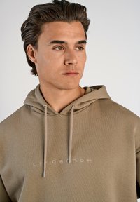 Sudadera beige con acabado texturizado, que cuenta con una capucha con cordón y el logo bordado "LINDENBERGH" en un tono a juego.