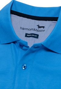 Polo blu realizzato in tessuto testurizzato con colletto, due bottoni e un collo interno a righe. Logo e etichetta "REGULAR FIT" visibili.