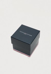 Tommy Hilfiger branded dark blue watch box with white, red, and blue stripes along the bottom edge on a plain light surface.