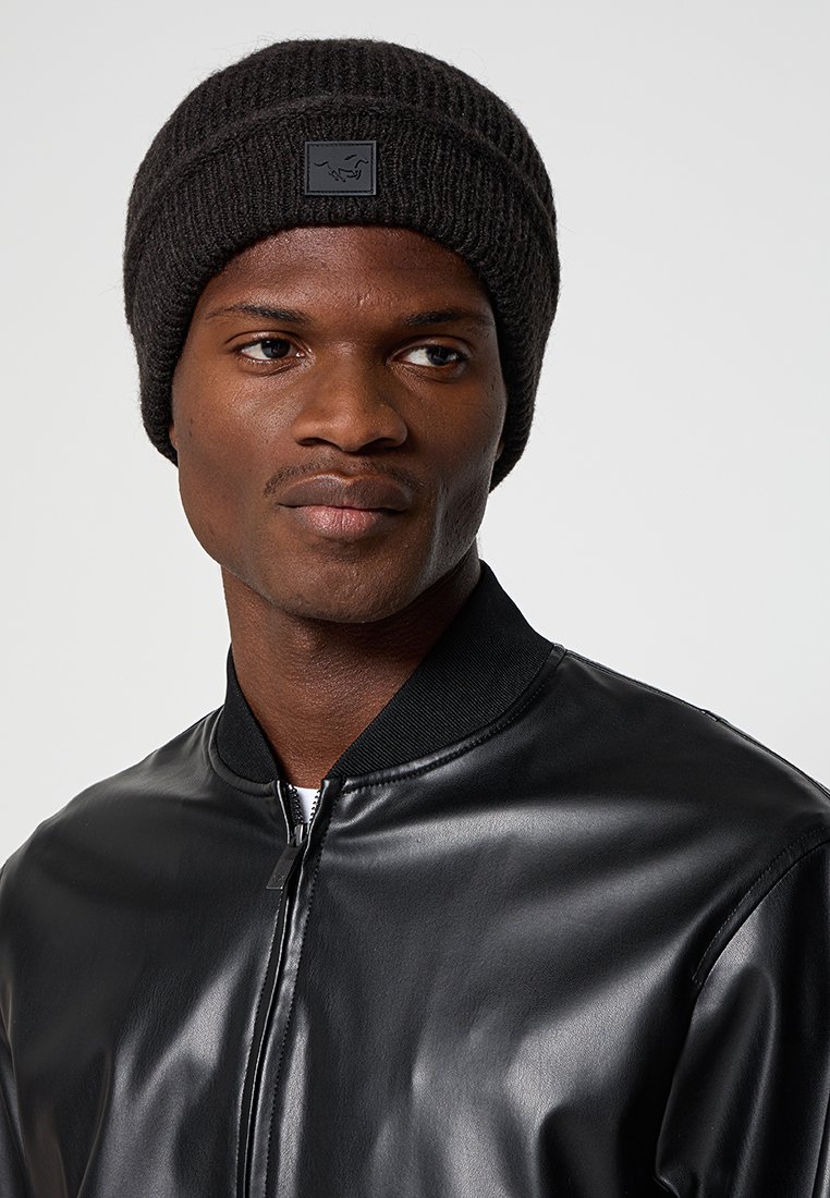 Czarna, prążkowana dzianinowa czapka z prostokątną łatą z logo, noszona z czarną skórzaną kurtką bombową. Zbliżenie na detale czapki i kurtki.