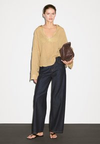 Maglione beige a coste, morbido, con scollatura a V, abbinato a pantaloni larghi in denim scuro. La modella tiene una borsa intrecciata marrone. Infradito neri.