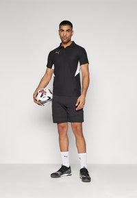 Svart polo-shirt med hvite detaljer, kombinert med svarte shorts og svarte joggesko. Holder en fotball. Enkelt design, funksjonelle materialer.