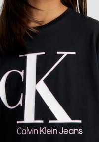 Černé bavlněné tričko s velkým bílým a růžovým logem "CK" a textem "Calvin Klein Jeans" pod ním v růžové barvě, kulatý výstřih.