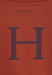 Červená tkanina s veľkým tmavomodrým písmenom "H" potlačeným pod textom "HACKETT" pri zaoblenom šve okolo krku.