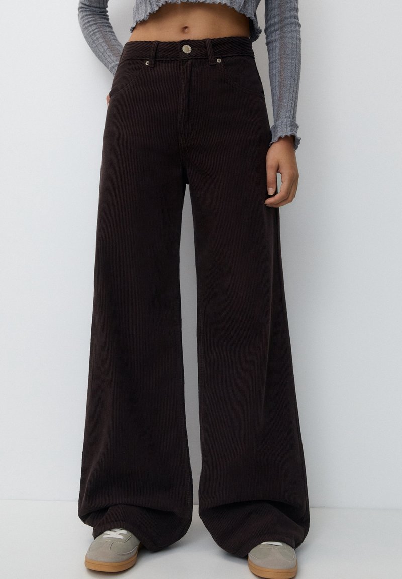 Culotte corduroy broeken in donkerbruin, met een gestructureerde afwerking, hoge taille en twee voorzakken. In combinatie met grijze sneakers.