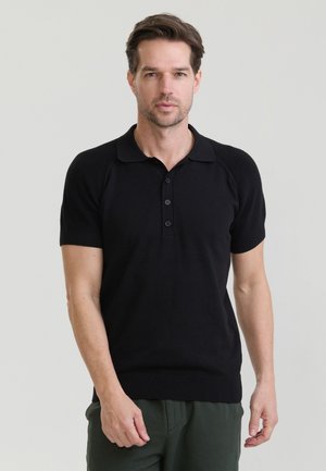 Man met kort bruin haar, gekleed in een zwart polohemd met korte mouwen en donkergroene broek, staand voor een effen lichte achtergrond.