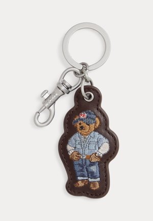 Võtmehoidja metallrõnga ja konksuga, millel on tumepruun nahkmärk, millel on tikitud karu sinist mütsi, särgi ja teksadega.