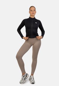 Zwarte sportjack met ritssluiting en een getailleerd ontwerp, gecombineerd met tan leggings met contrasterende naden en een logo. Witte sportschoenen maken de look af.